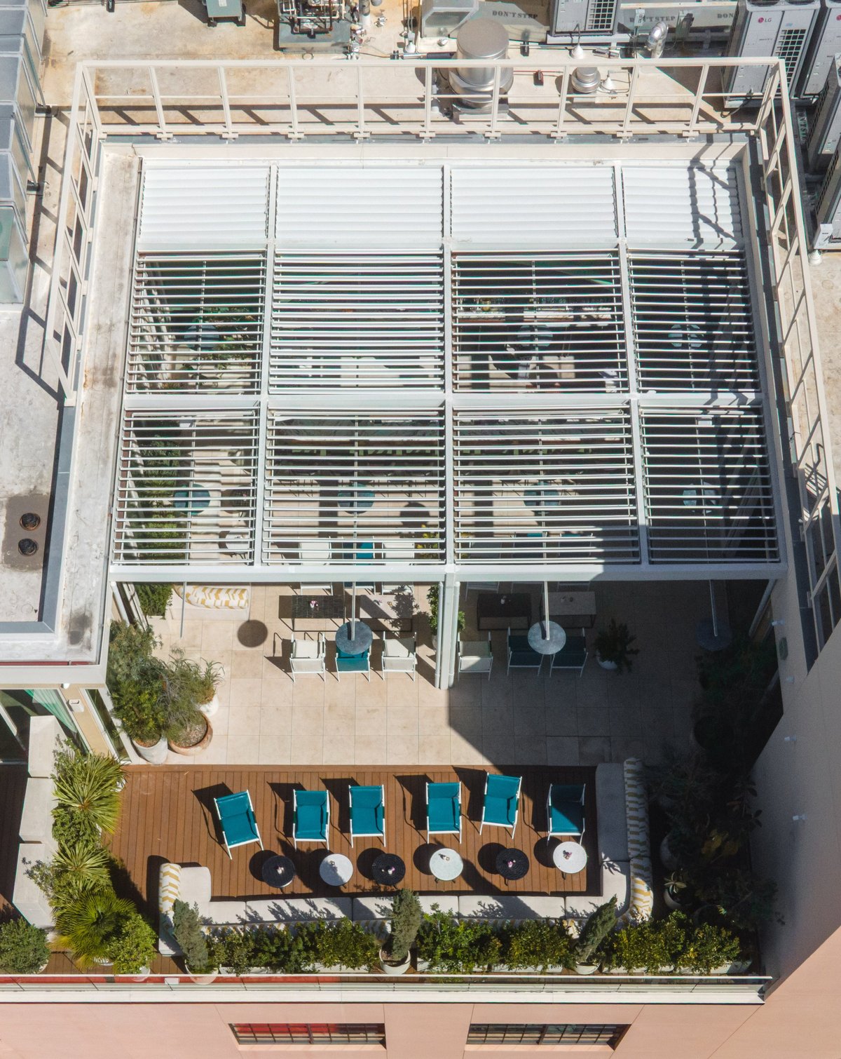 Aerial view of a modern rooftop with wooden deck, lounge chairs, and large white louver screens shading the upper level