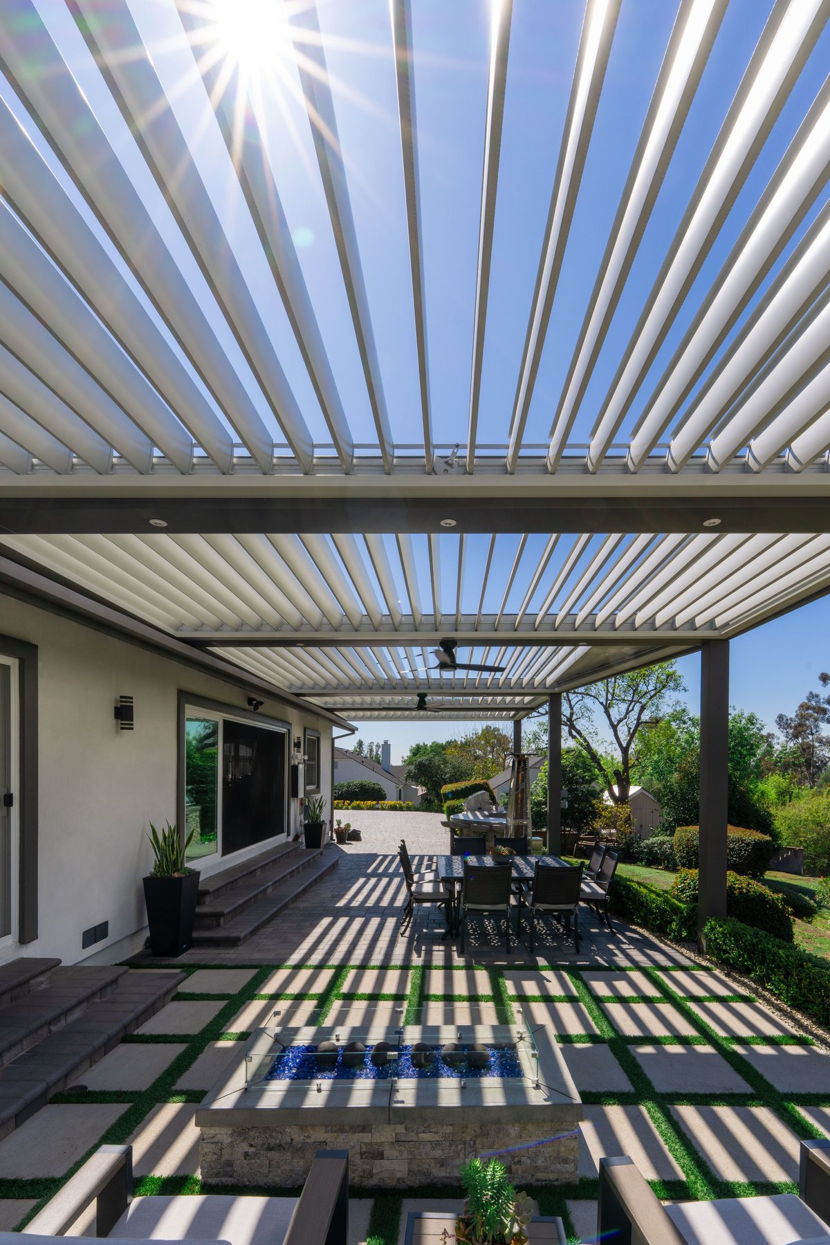 Modern patio with louvered pergola roof, dining furniture, and landscaped garden under bright sunlight