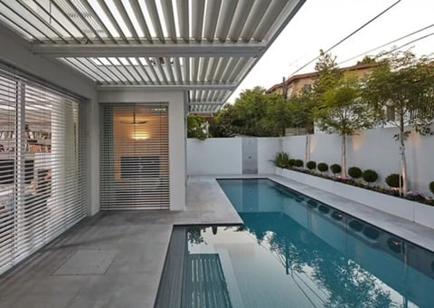 Modern backyard with narrow lap pool, louvered pergola, gray patio, and white boundary fence with landscaping