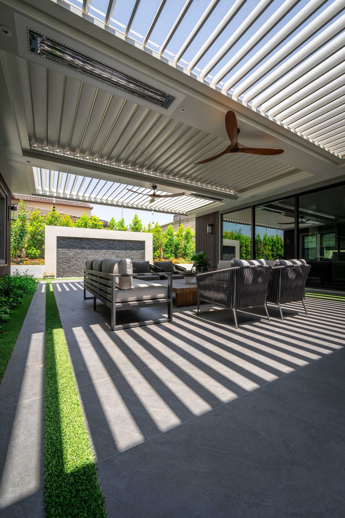 Modern outdoor patio with louvered pergola, metal lounge chairs, and striped shadows cast across the concrete floor