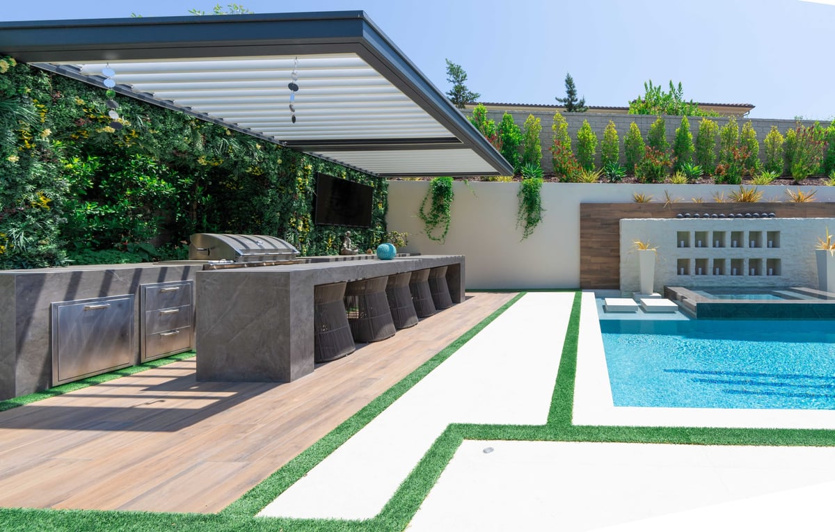 Modern backyard patio with swimming pool, retractable awning, dining table, and wooden deck surrounded by vegetation