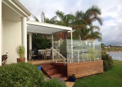 Modern waterfront home with wooden deck, glass-enclosed patio, metal pergola, and palm trees overlooking a scenic water view