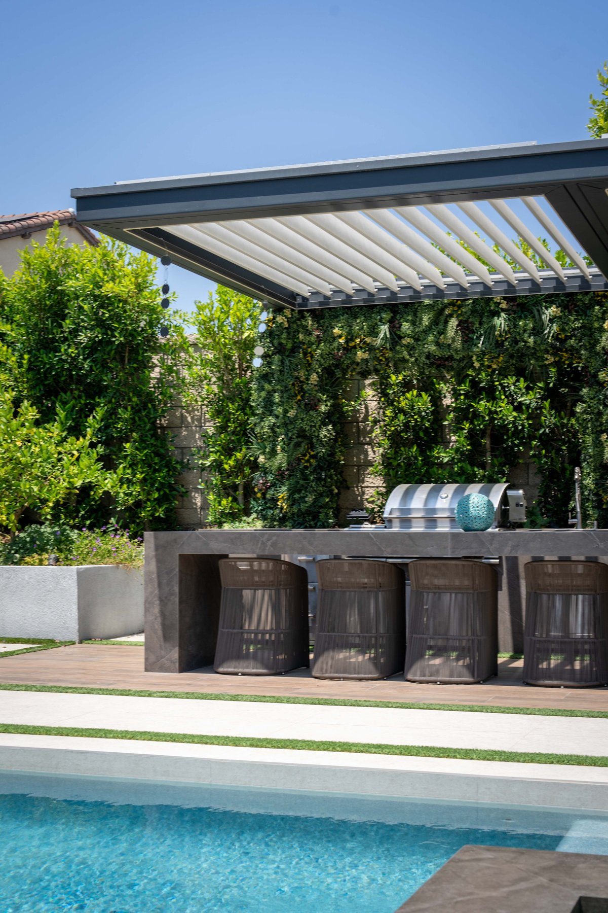 Modern pergola with retractable louvered roof over outdoor kitchen with stainless steel grill, next to swimming pool